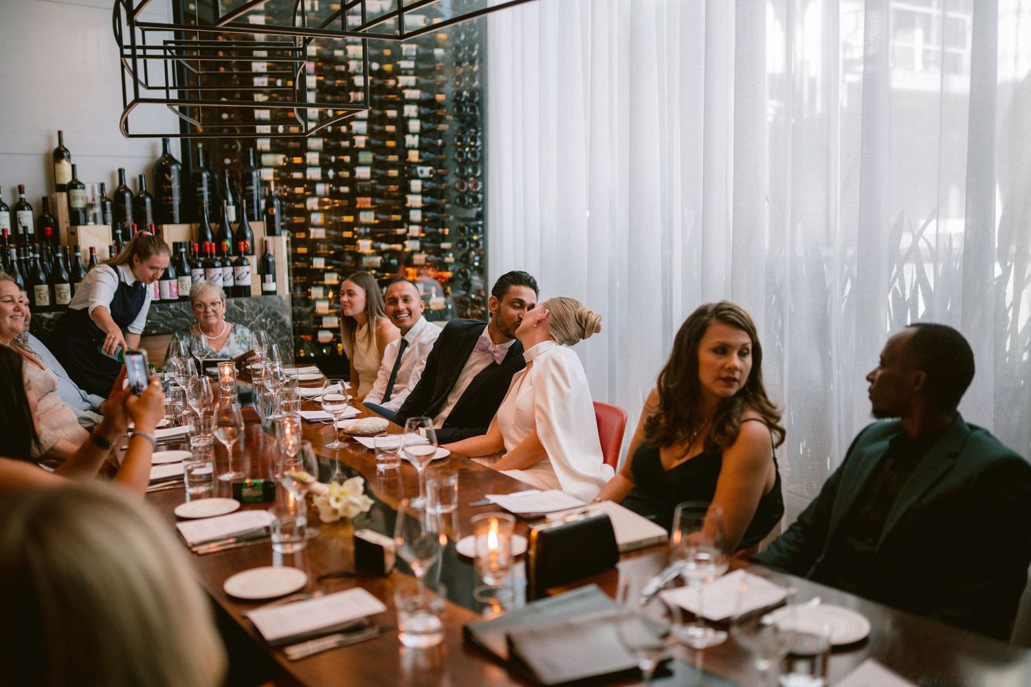Wedding reception guests seated at long dining table in upscale restaurant with wine wall and candlelight ambiance.