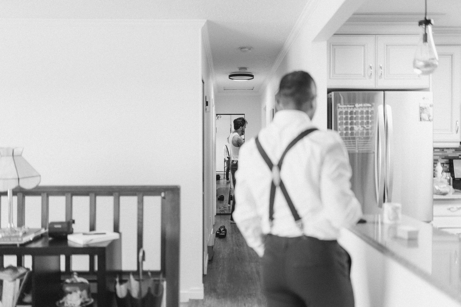 Black and white photo of groom in suspenders walking through home hallway on wedding day.