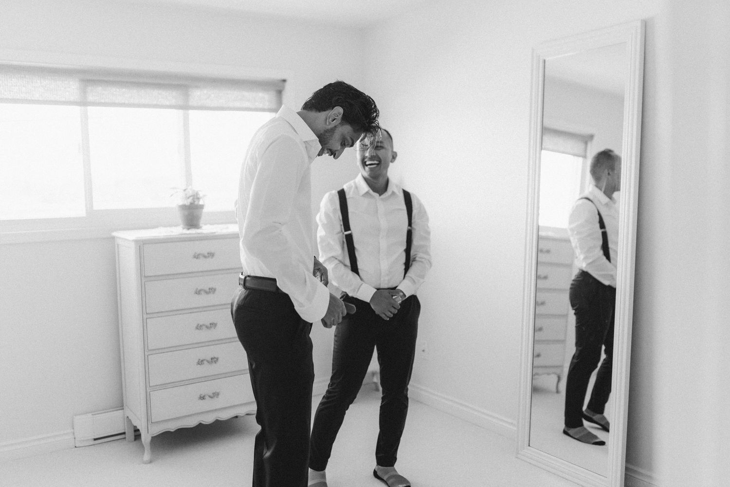 Groom and groomsman laugh together while getting ready in white room with mirror reflection on wedding day.
