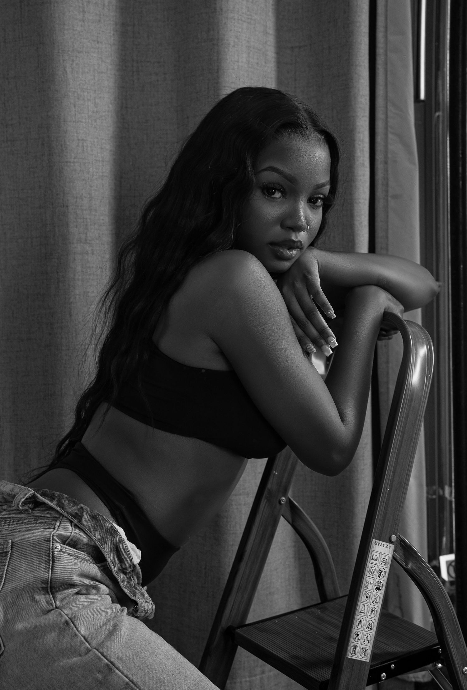 Black and white photo of woman in crop top sitting on chair by window.