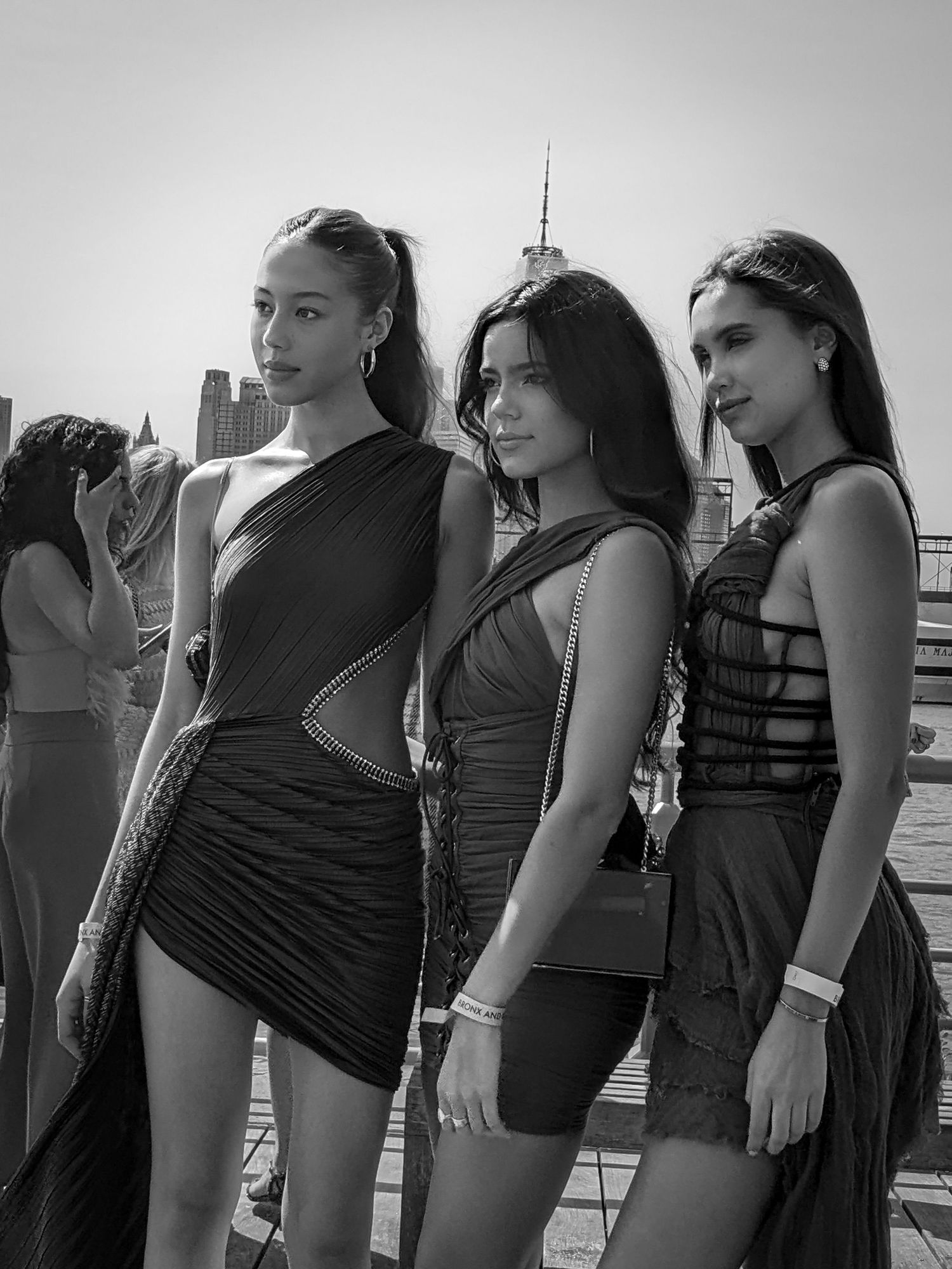 Three women in black dresses posing on rooftop with city skyline behind.