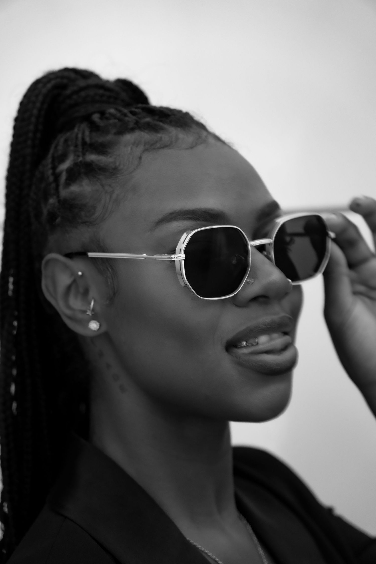 Close-up black and white portrait of a woman with braided hair adjusting geometric silver sunglasses.