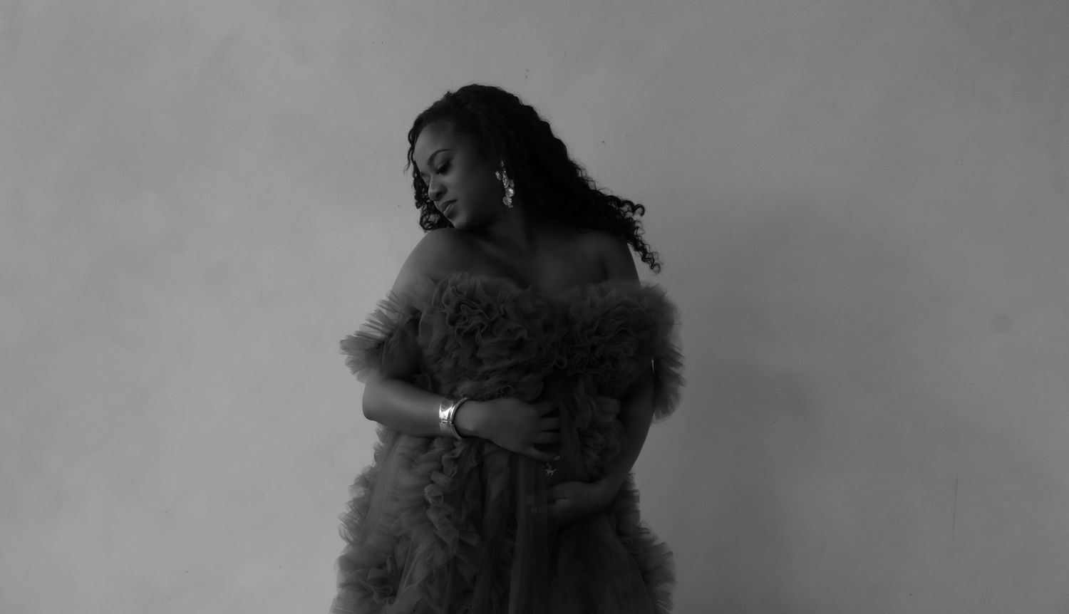 Black and white artistic portrait of a woman in a fluffy off-shoulder gown embracing herself against a soft background.