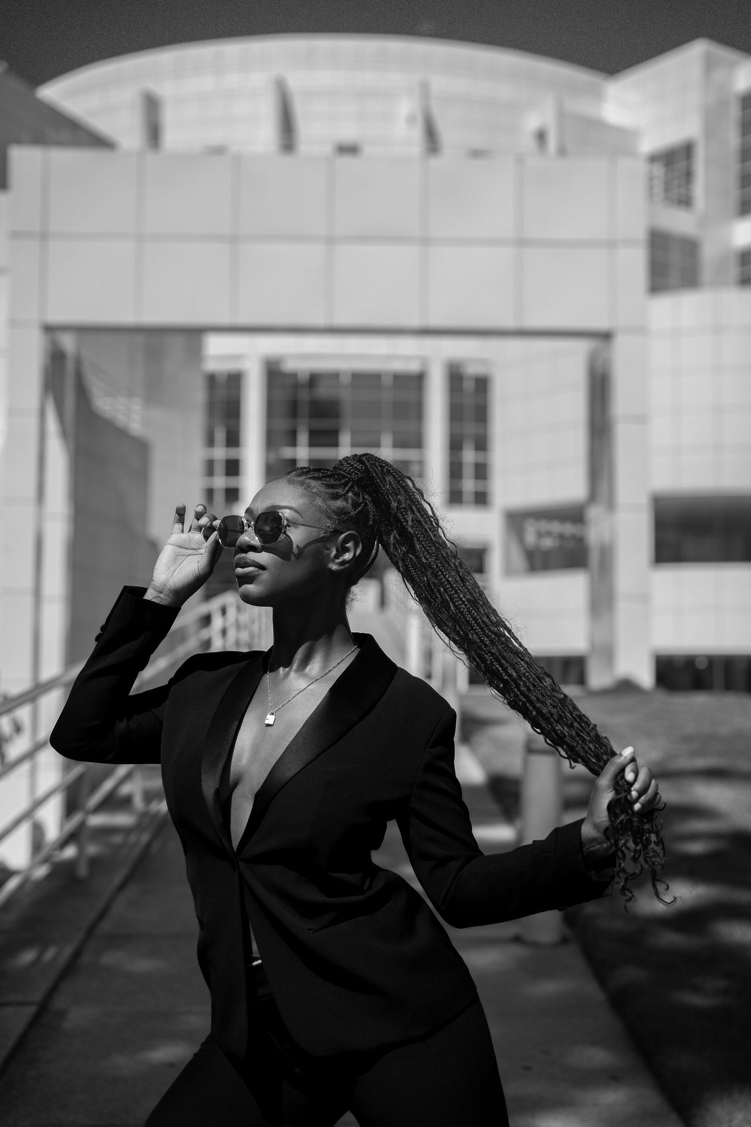 Black and white photo of a woman in a black suit adjusting sunglasses outdoors near a building.