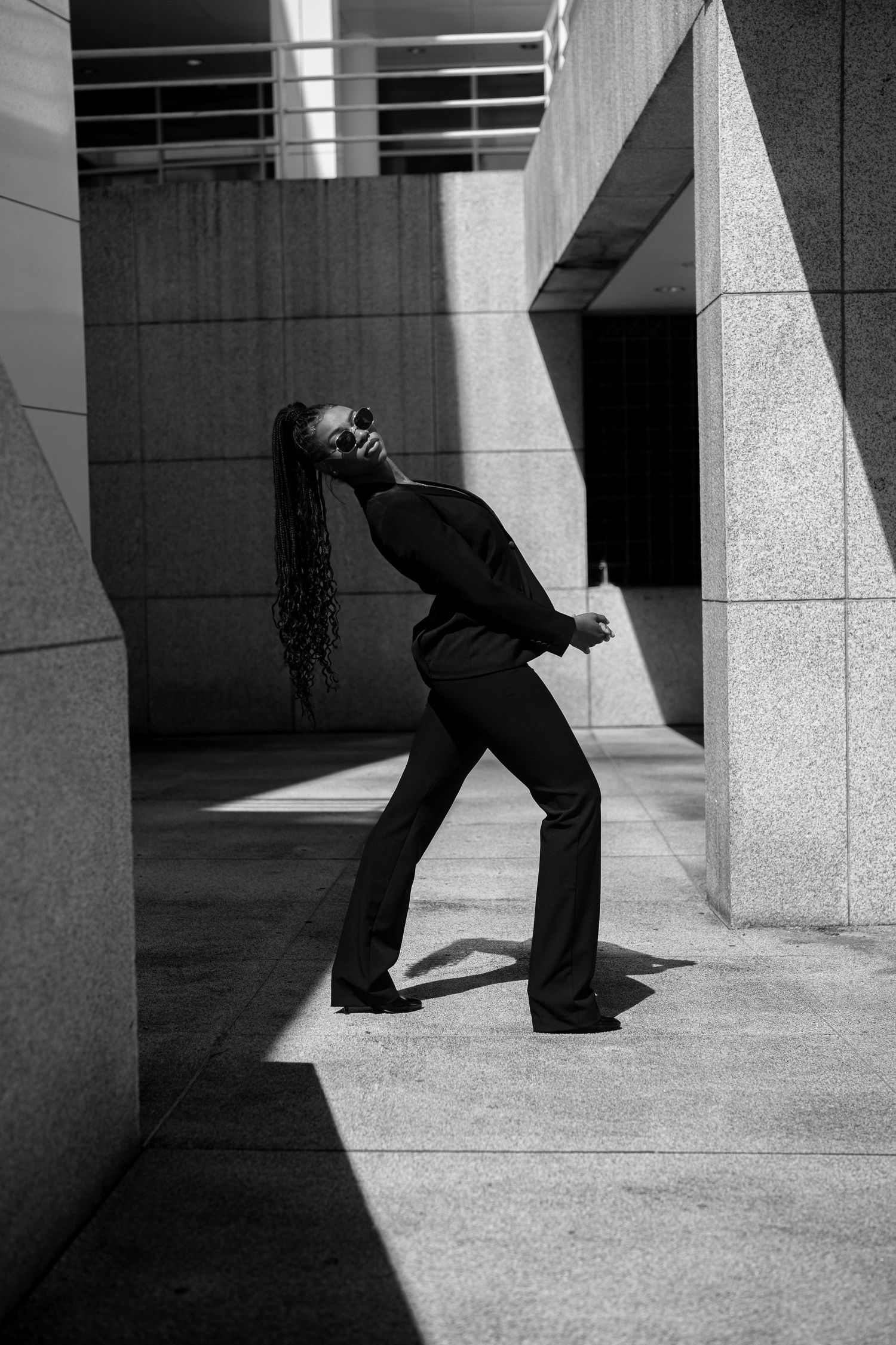 Black and white photo of a woman in a black pantsuit posing dramatically between concrete columns.