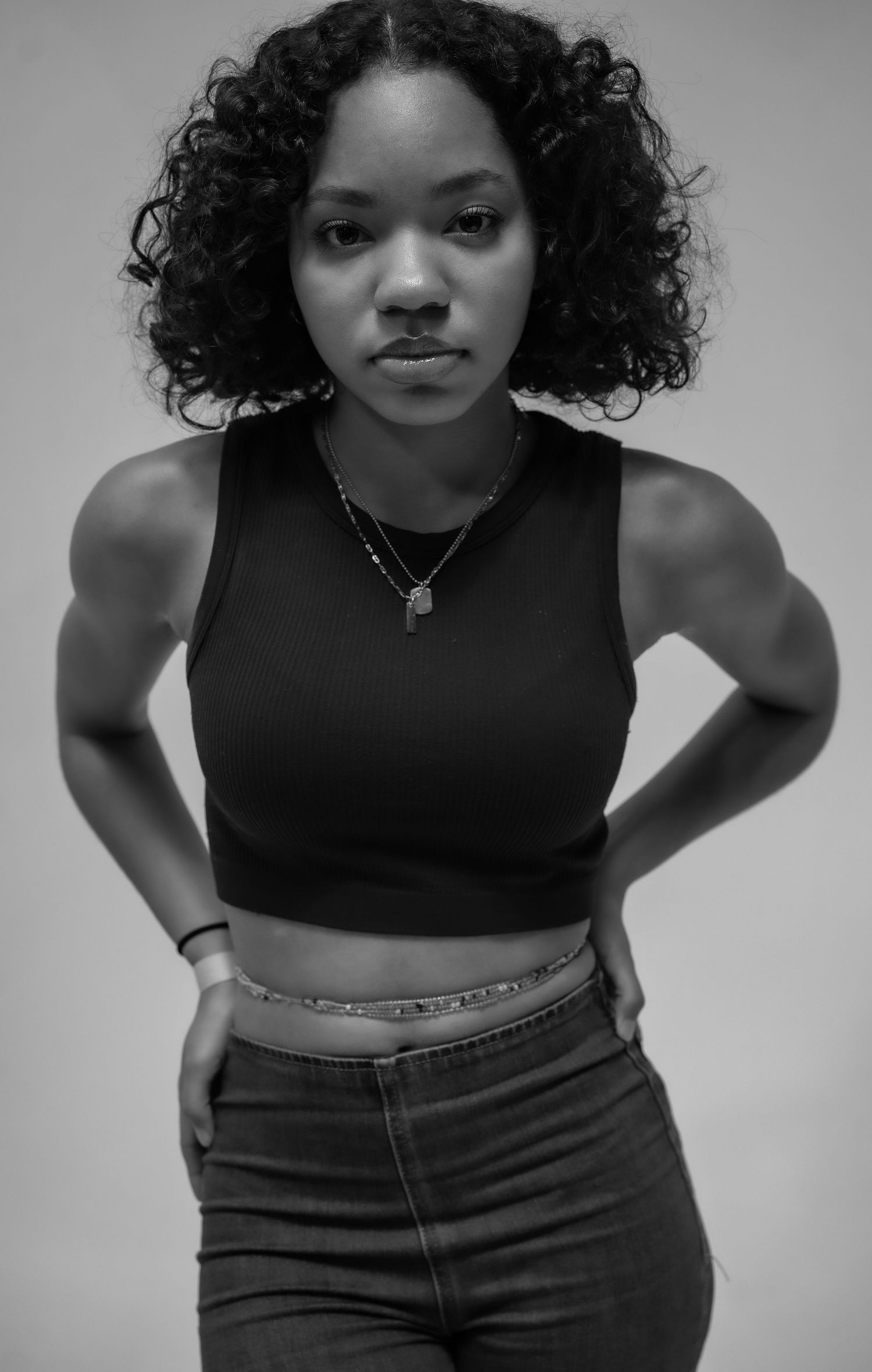 Black and white portrait of a young woman with curly hair wearing a crop top and jeans, posing confidently.
