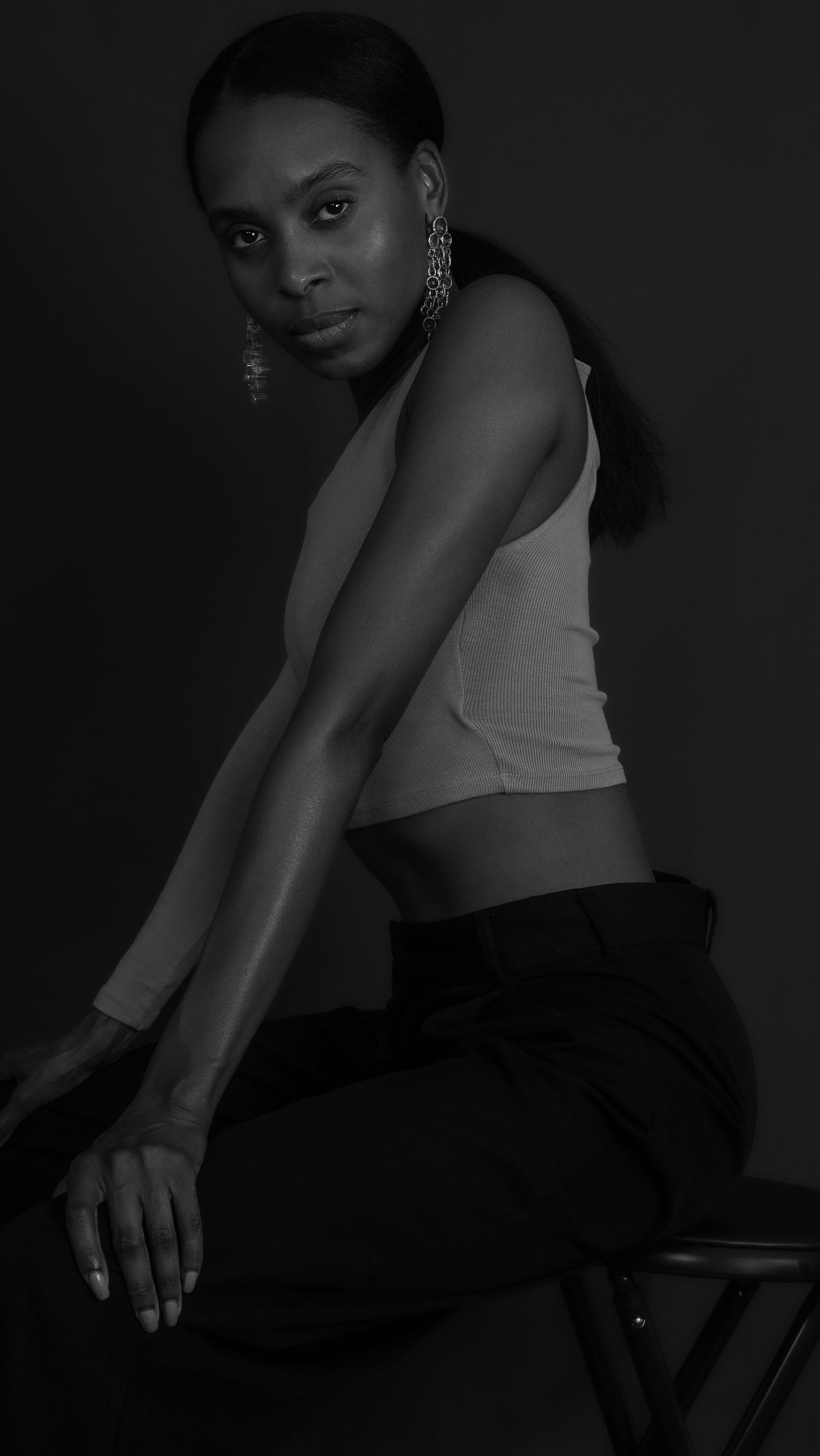 Black and white studio portrait of a woman in a crop top and black pants seated on a stool.