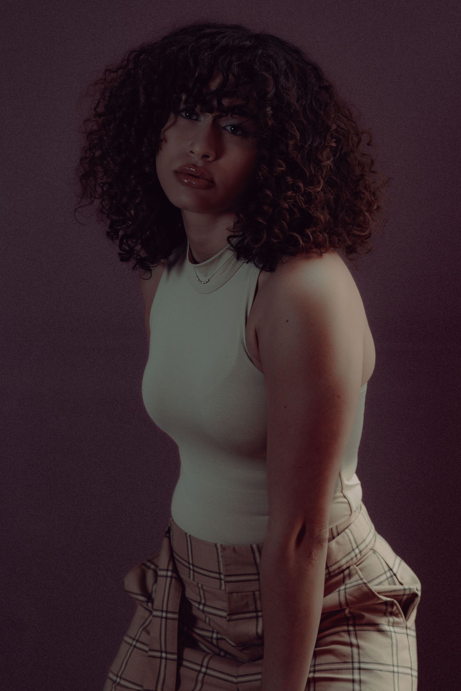 Young woman with curly hair wearing a mint sleeveless top and plaid pants posing against a dark background.