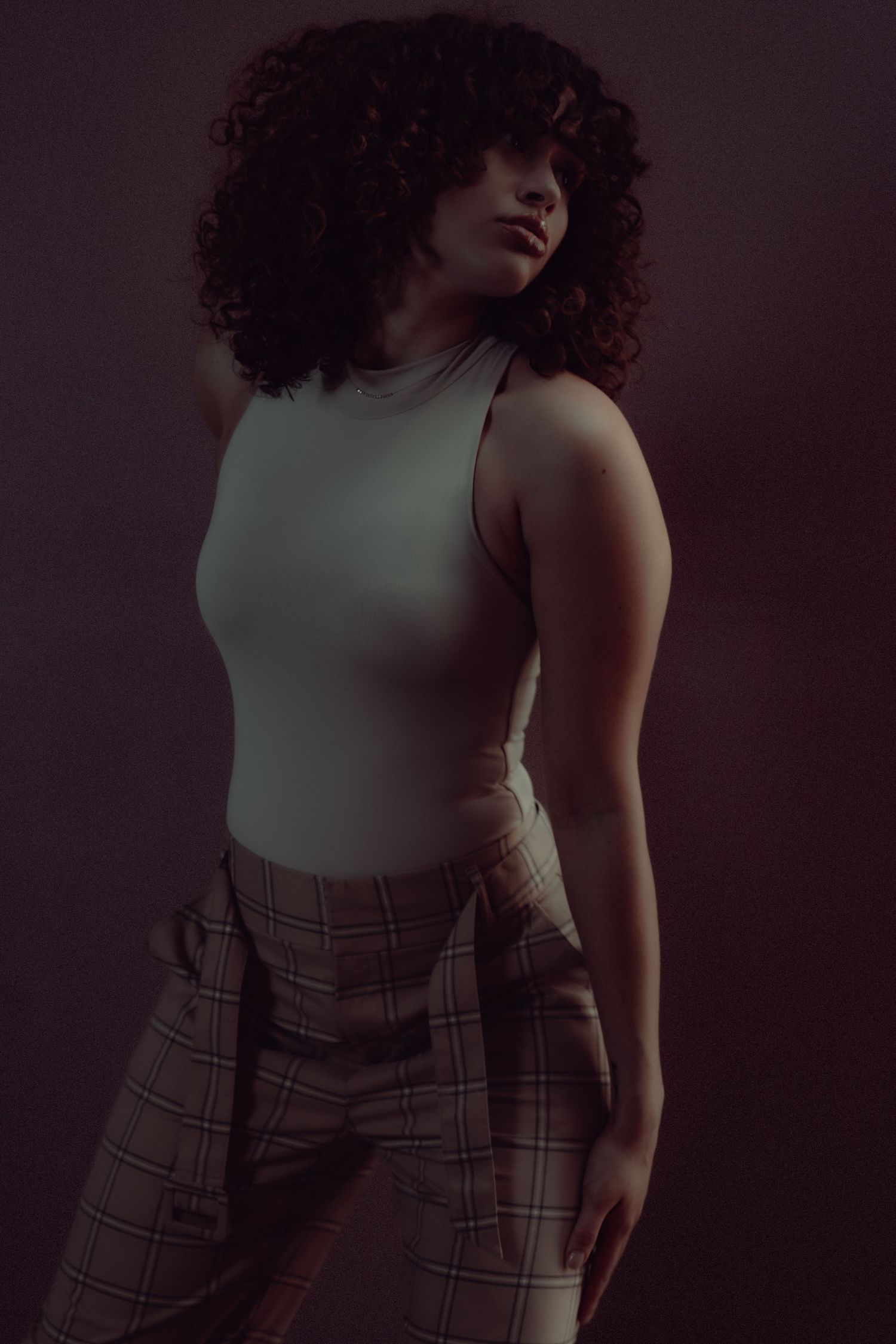 A young woman with curly hair poses in a white sleeveless bodysuit and plaid trousers against a dark background.