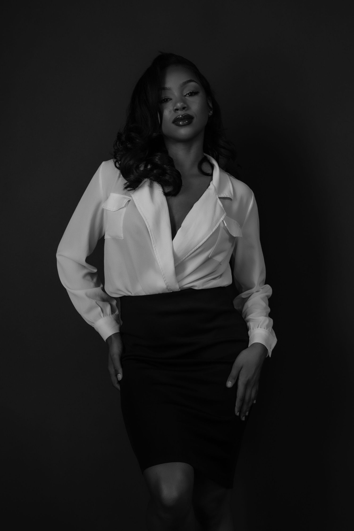 Black and white portrait of a woman in a white blouse and black pencil skirt against dark background.