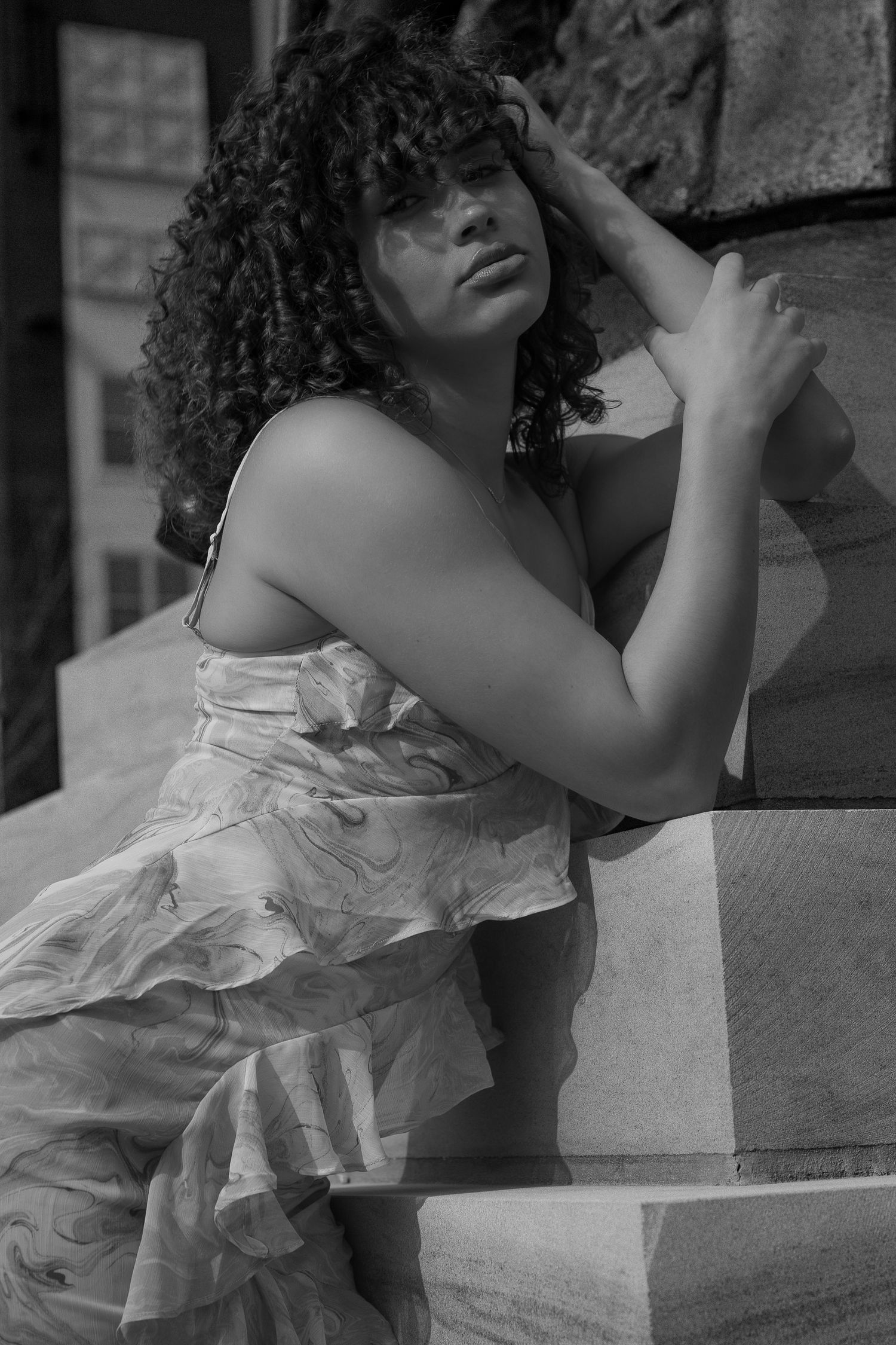 Black and white photo of a woman in a floral dress posing against a stone pillar with curly hair.