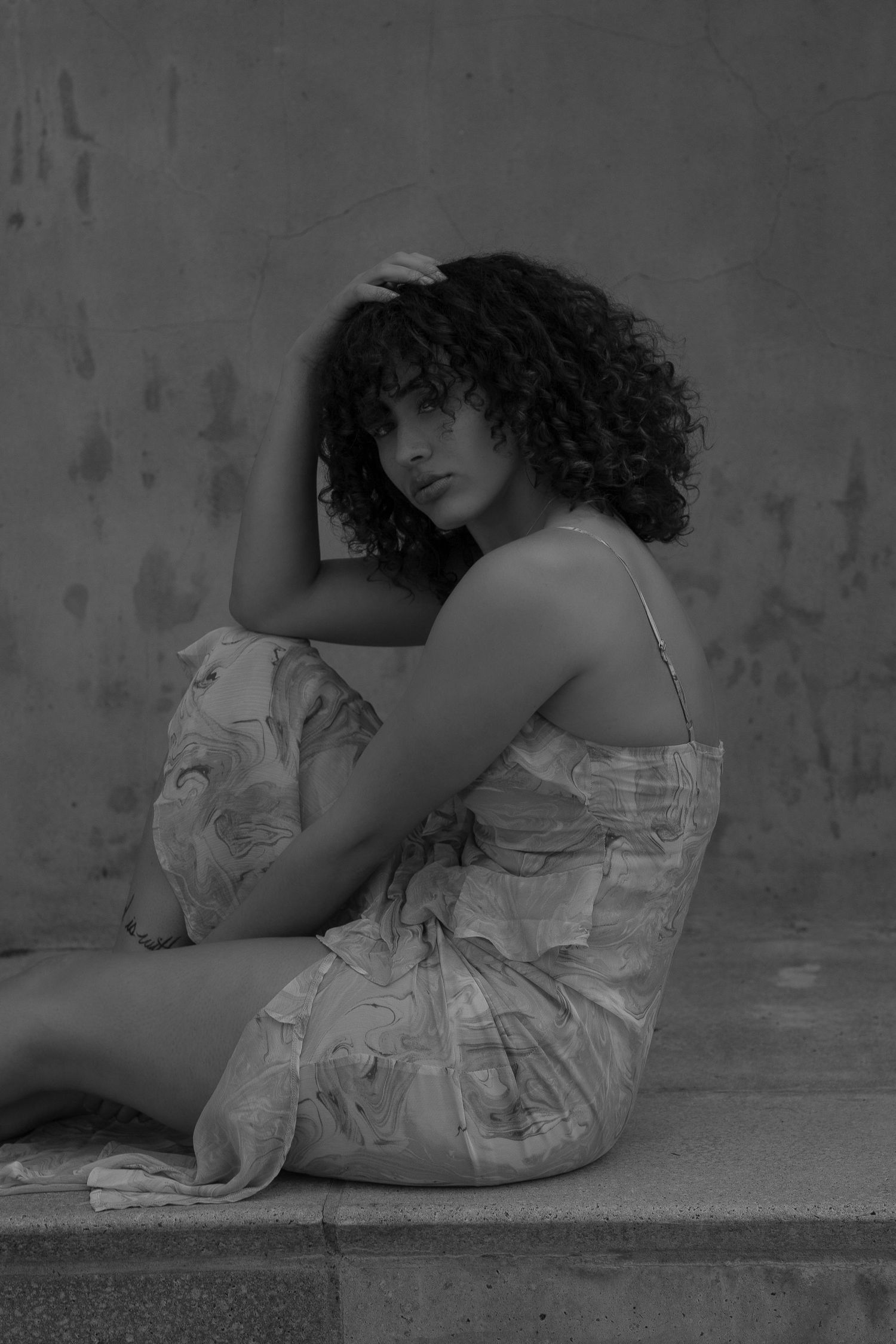 Black and white portrait of a young woman with curly hair in a floral dress, sitting against a textured wall.