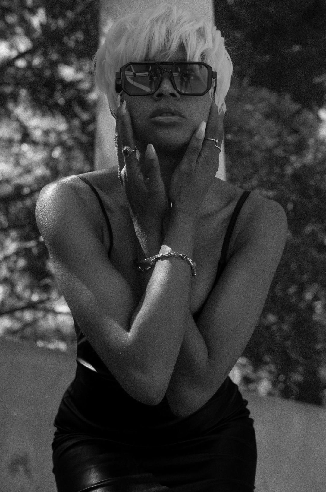 Black and white portrait of a woman in a black dress with blonde hair and oversized sunglasses posing outdoors.