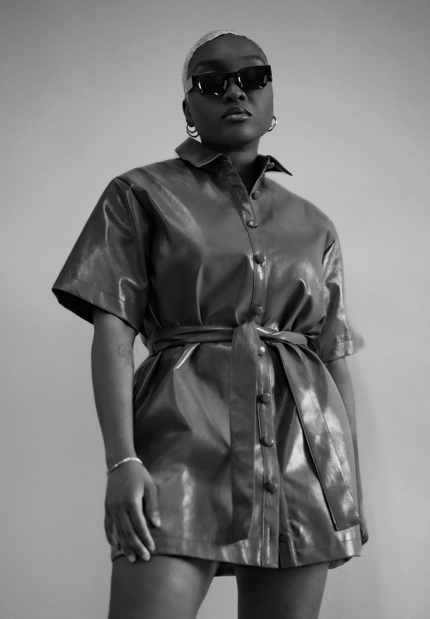 Woman in black leather belted shirt dress wearing sunglasses in B&W photo.