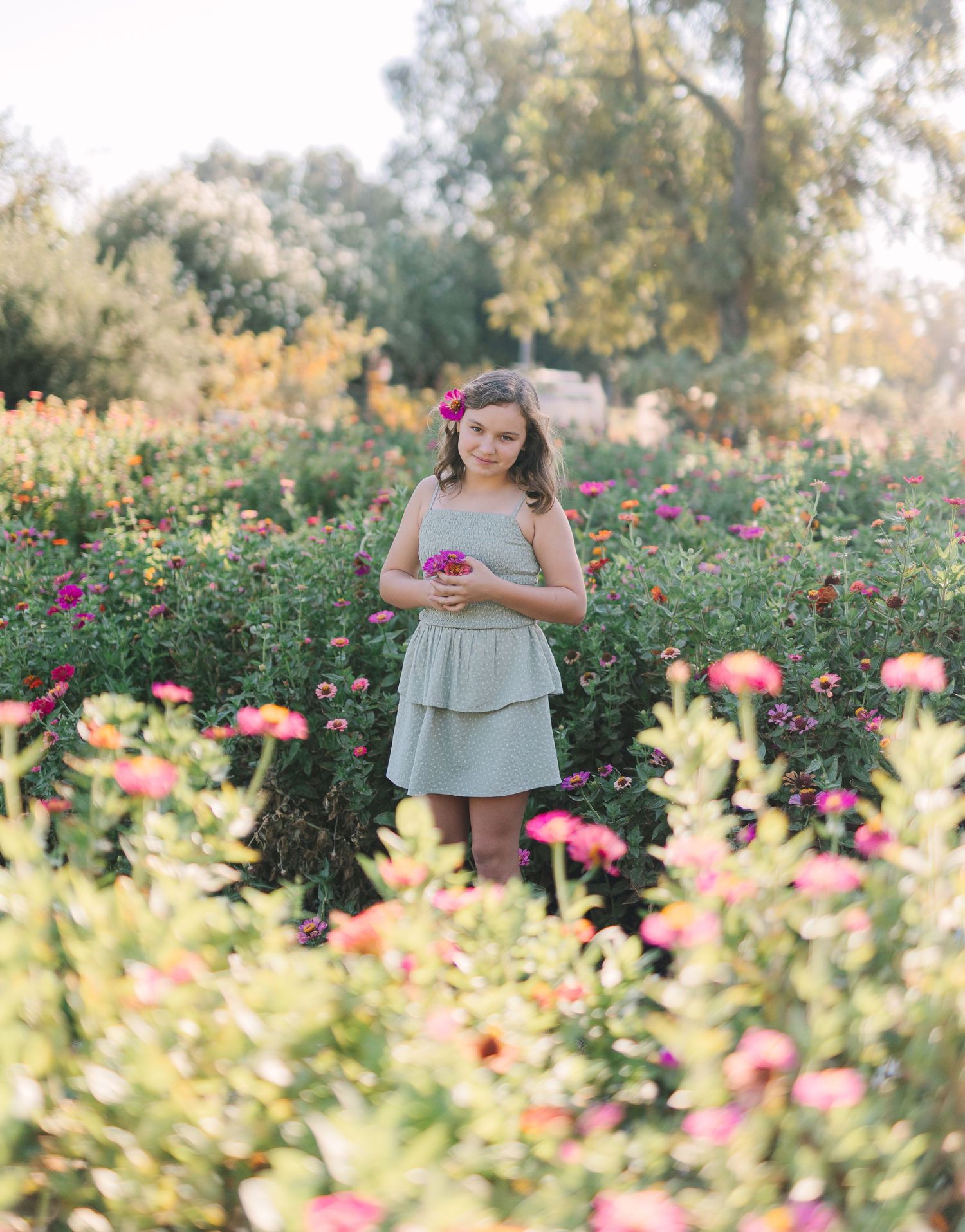 Young girl in dress holding pink flowers, standing in a colorful zinnia garden in soft natural light during a fall mini session with Jessica