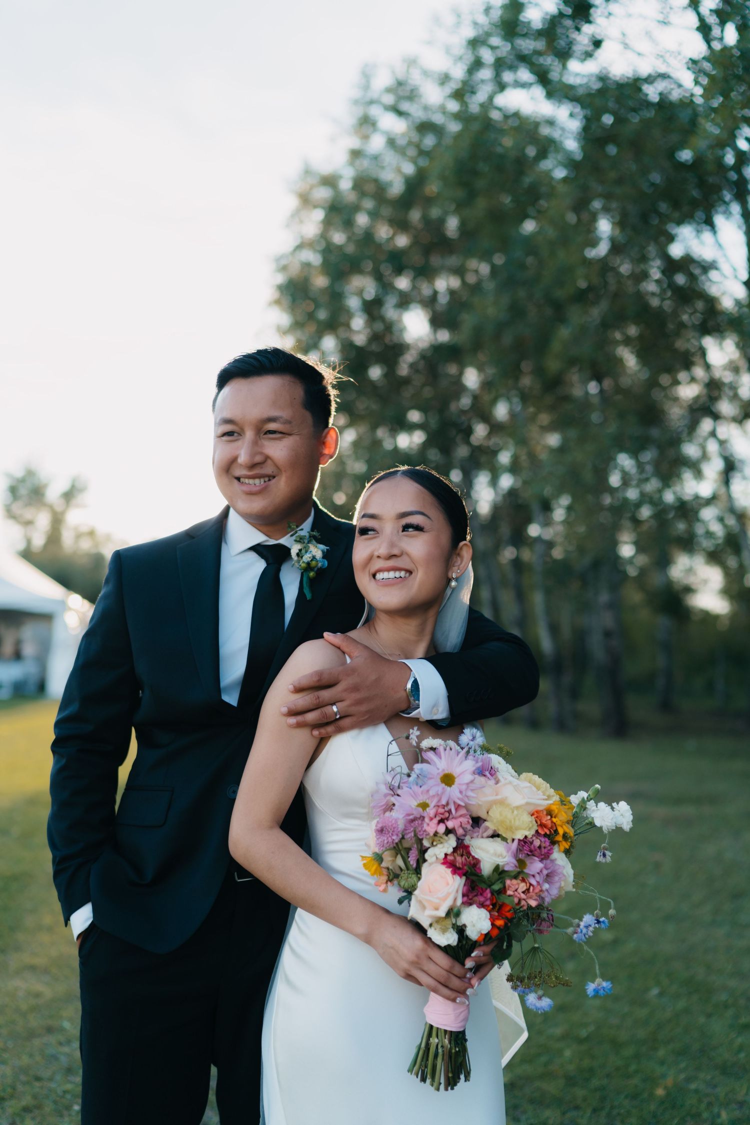 Bride and groom pose outdoors on green lawn, she holds colorful bouquet in white gown, he wears black suit with boutonniere.