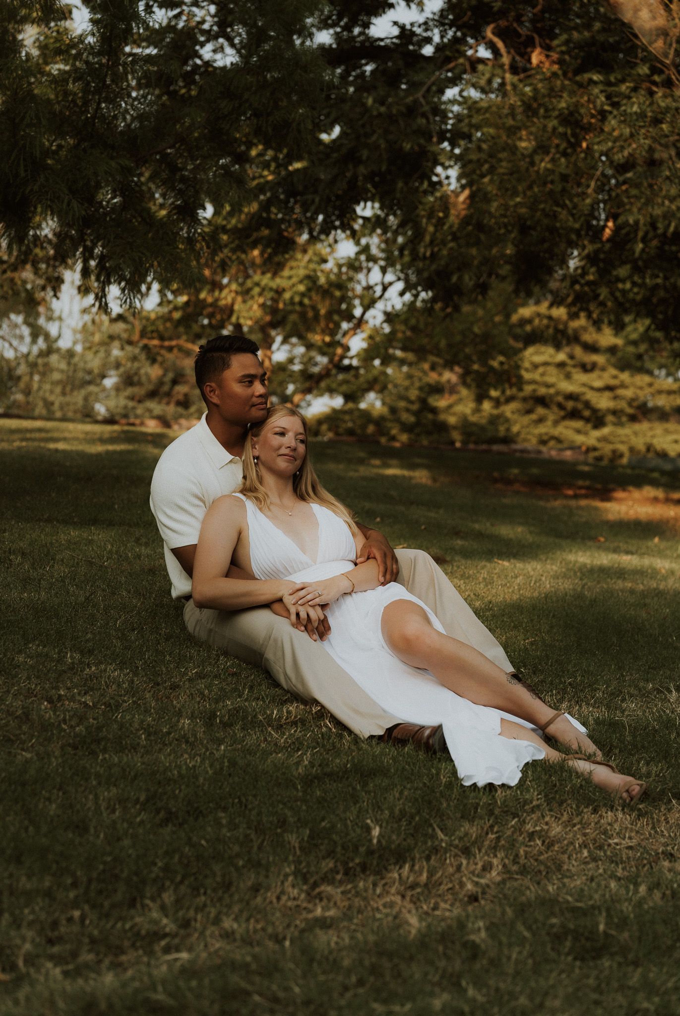 Couple sitting together on lush green grass under trees, woman in white dress leaning against man in white shirt.
