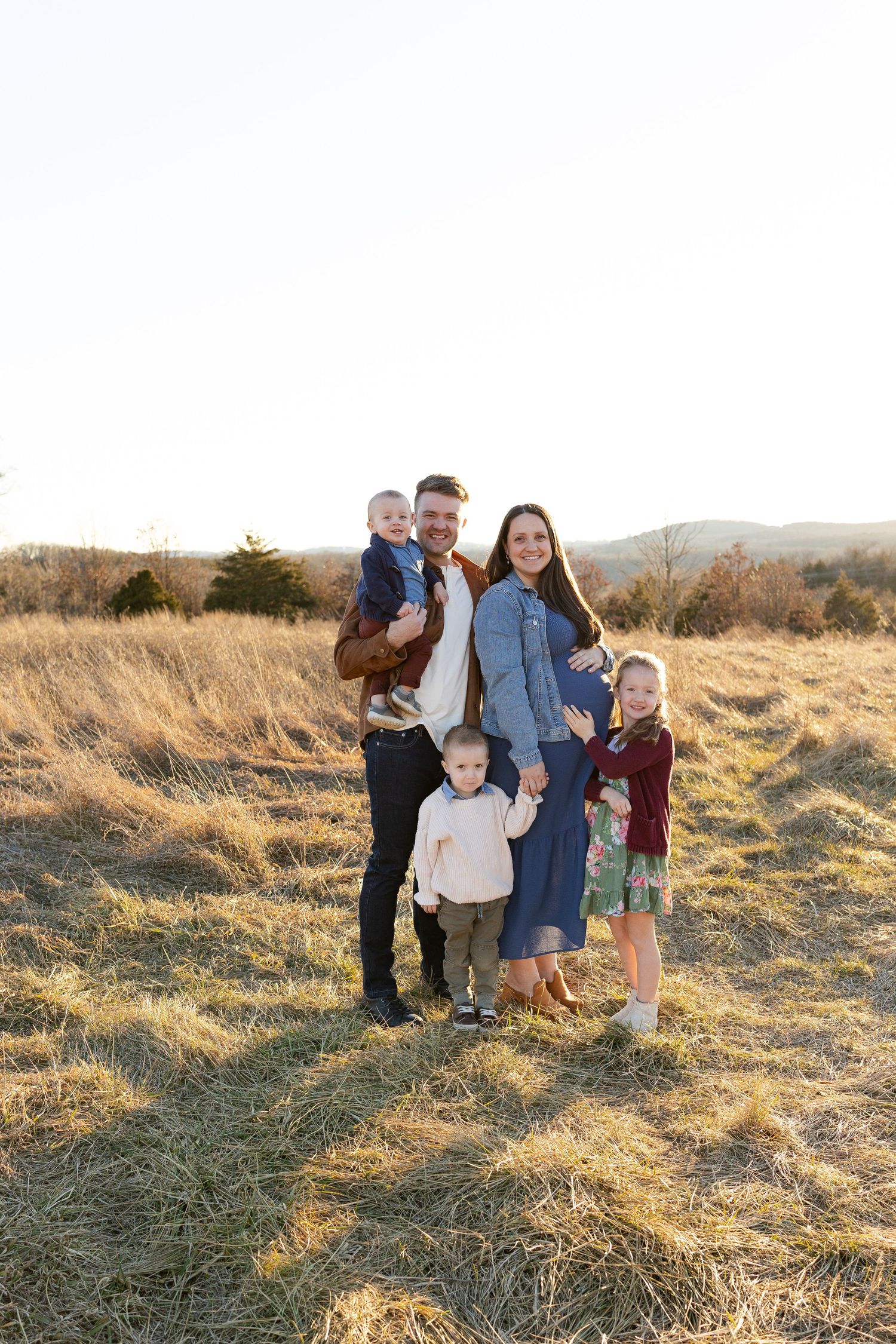 Family of five posing in golden field at sunset with mountain views in background.