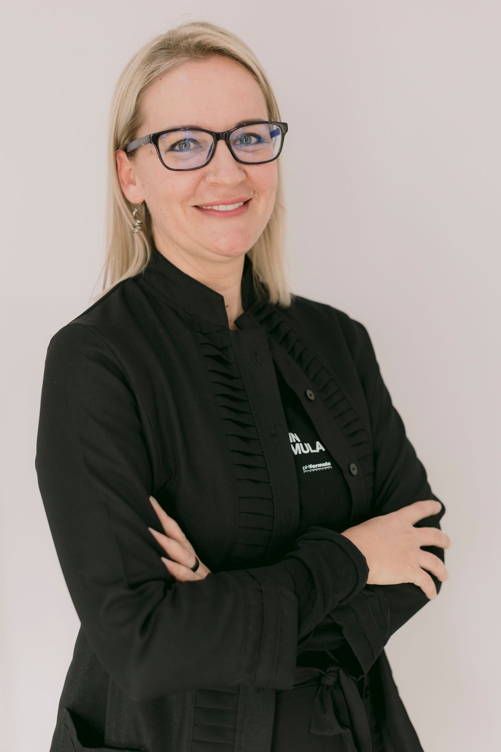 Smiling blonde woman with glasses in black chef uniform, arms crossed, professional portrait on light background.