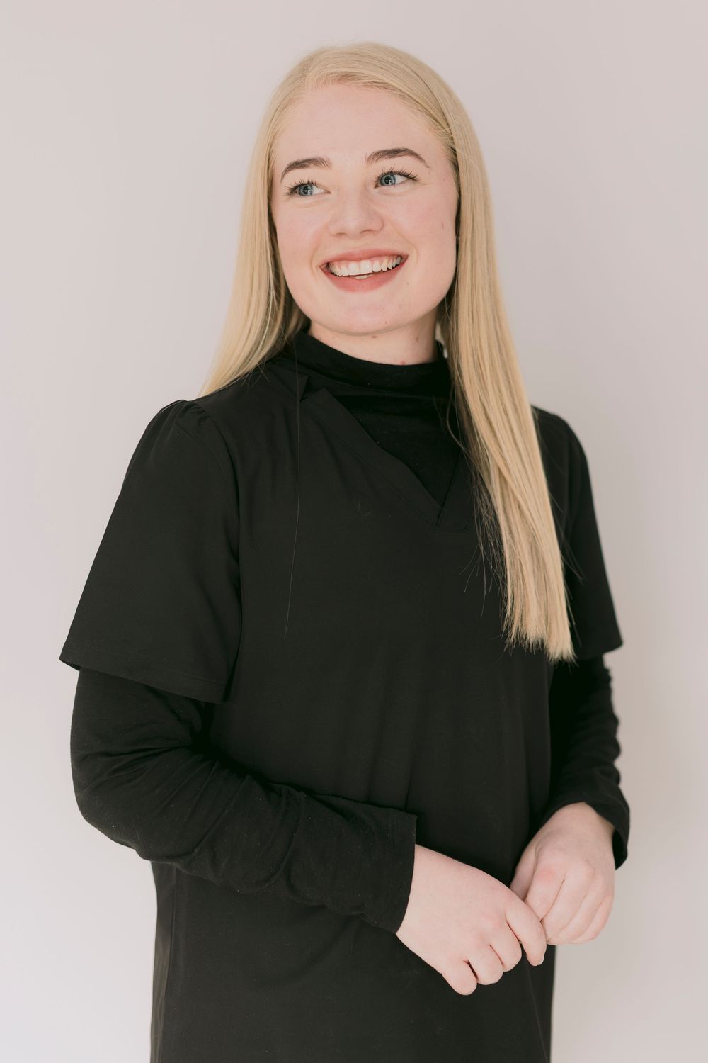 Smiling blonde woman in black turtleneck dress posing against light background.