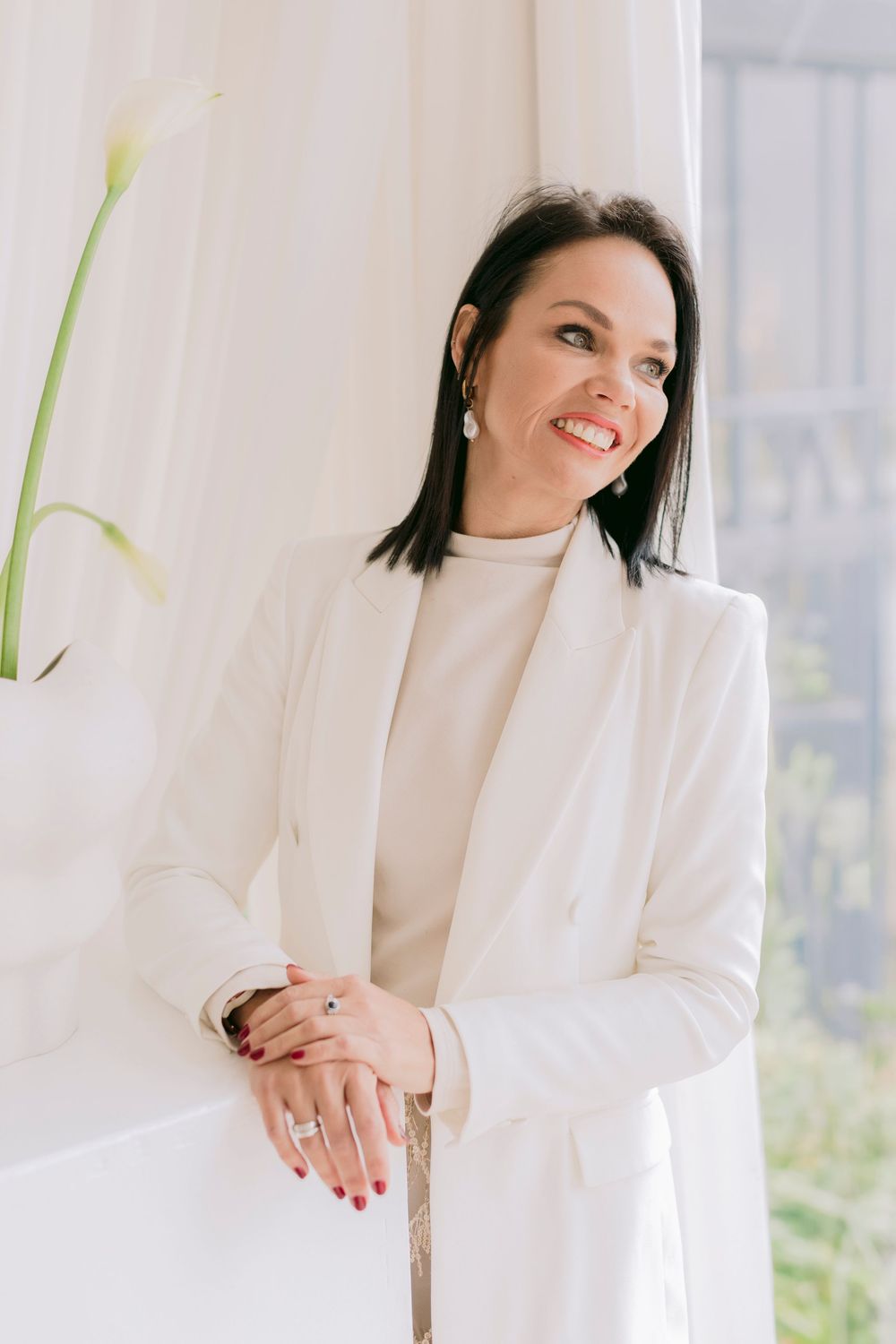 Smiling brunette woman in white blazer and dress poses elegantly beside white calla lilies near a bright window.