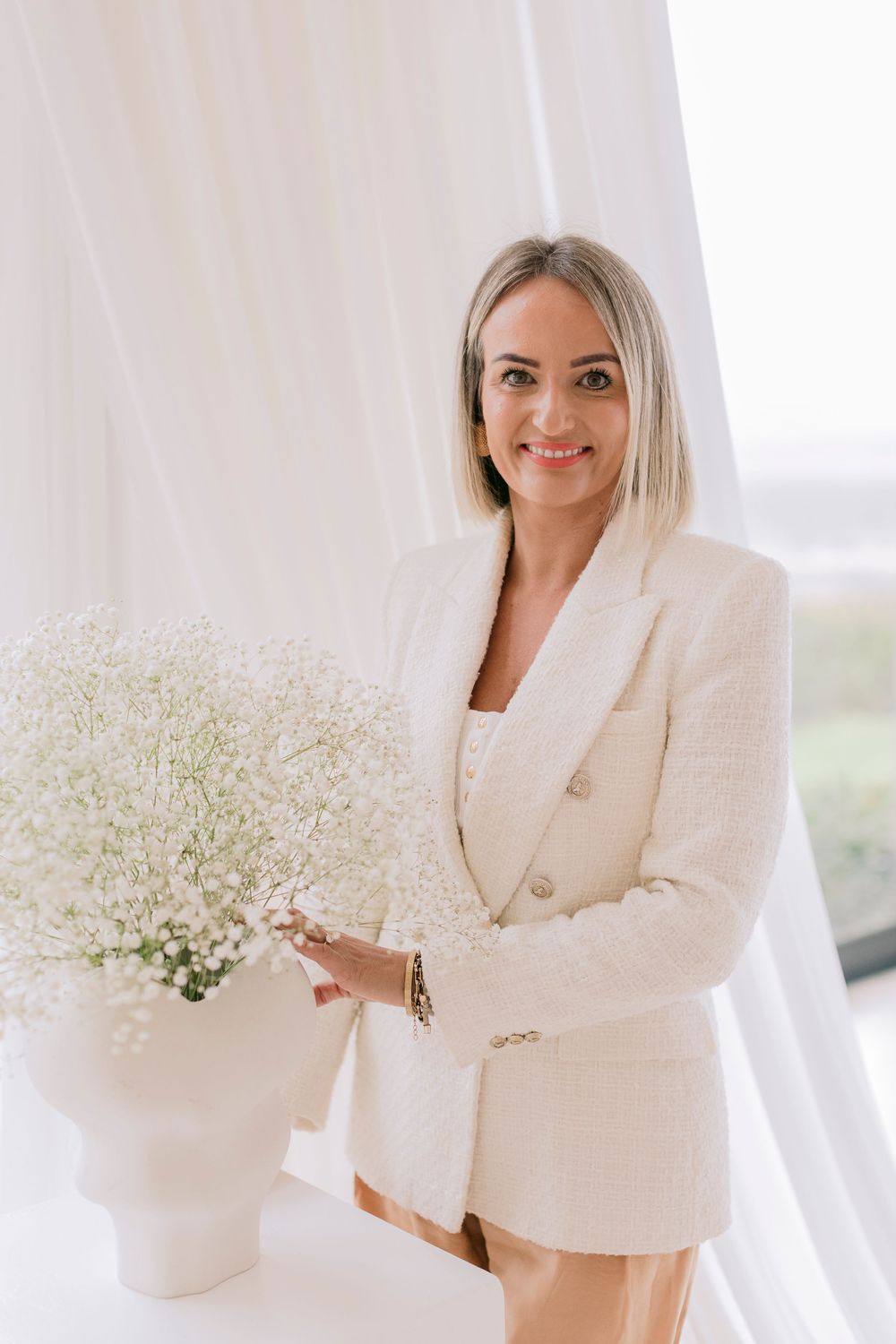 Smiling blonde woman in white blazer arranging baby's breath flowers in white vase near sheer curtains.