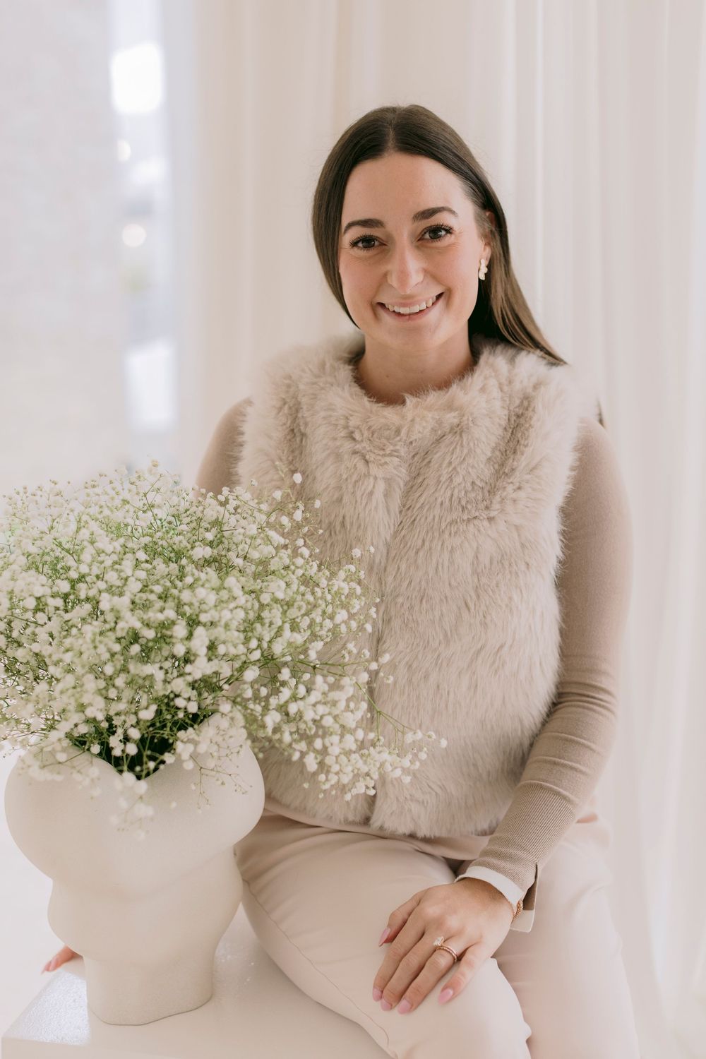 Smiling woman in a fuzzy vest sitting beside a white vase with baby's breath flowers in a bright room.