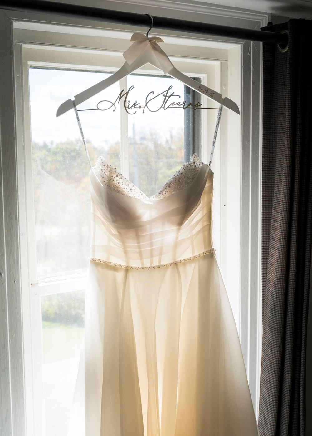 Wedding dress hanging on a personalized Mrs. Stearns hanger in front of a sunlit window.