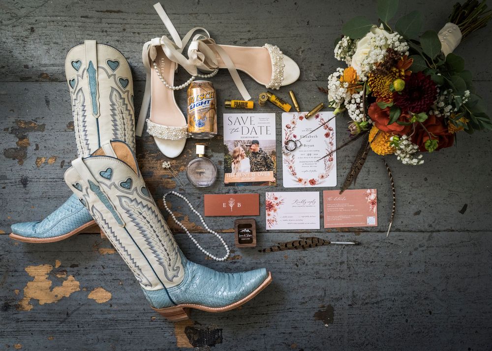 Blue cowboy boots, heels, bouquet, and wedding stationery flatlay on rustic wood floor.