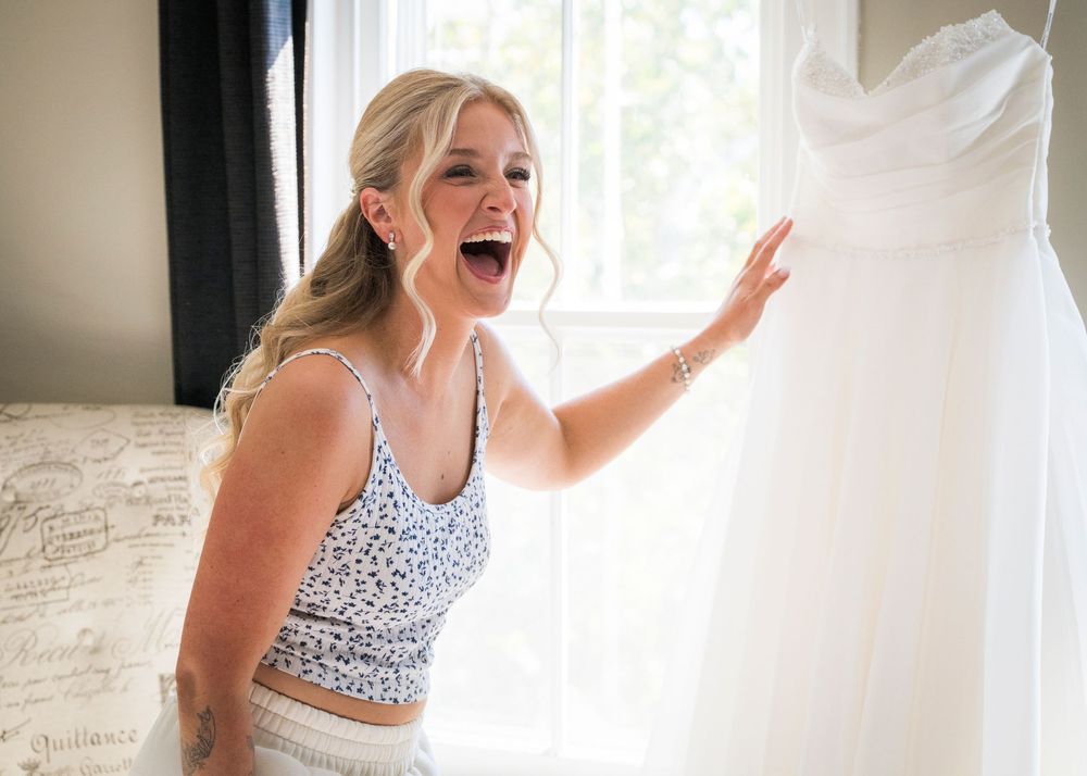 Excited blonde woman reacts with joy upon seeing her wedding dress hanging by window.