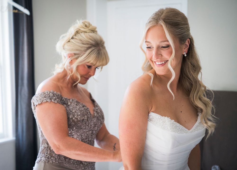 A smiling bride in a white gown gets help with her dress from a woman in a silver beaded gown.