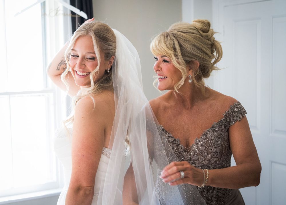 Smiling bride in white gown and veil with mother in beaded dress by bright window.