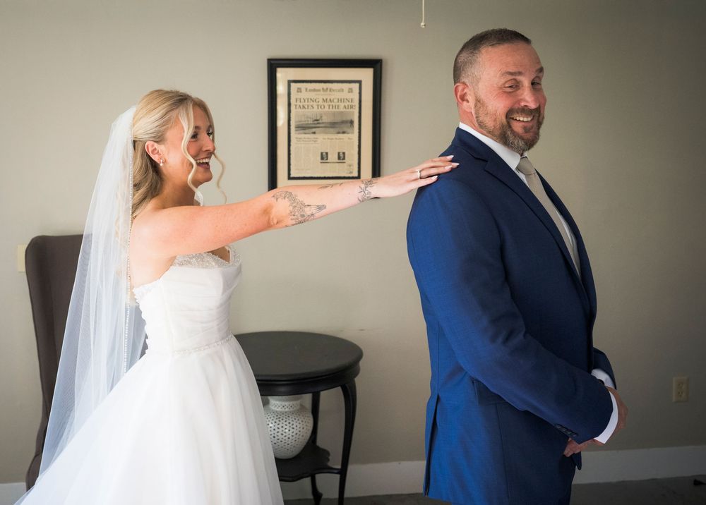 Bride in white gown taps groom's shoulder during first look moment indoors.