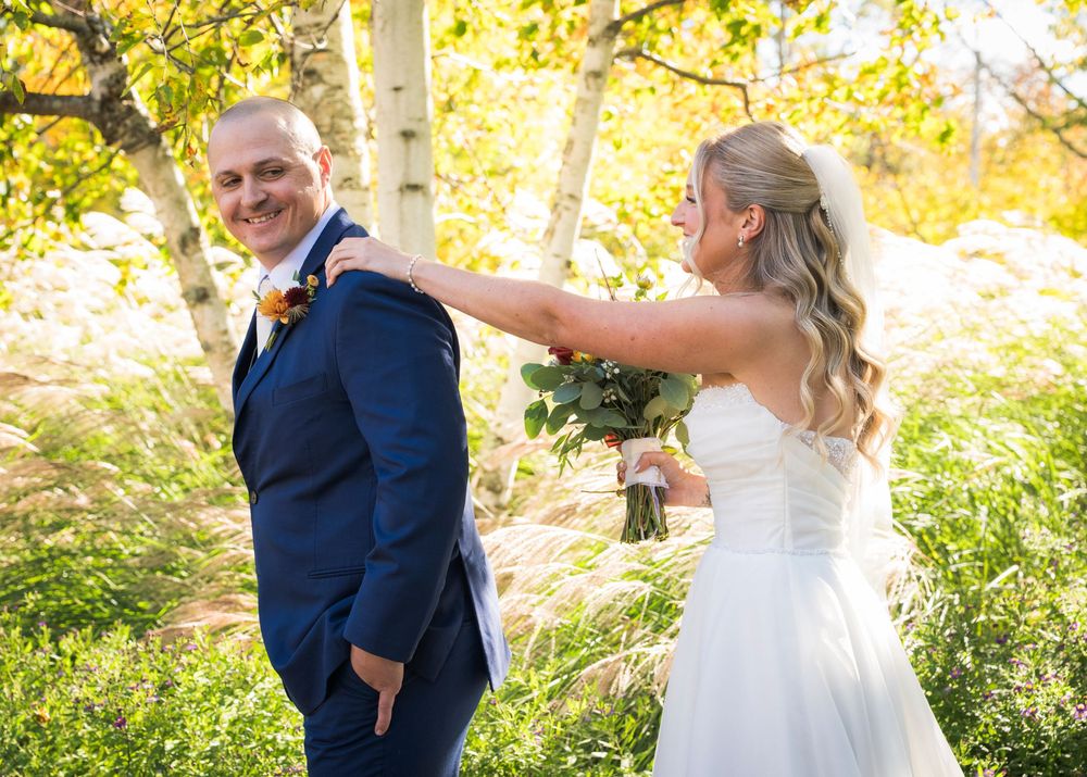 Groom in navy suit smiles as bride in white gown taps his shoulder for first look.