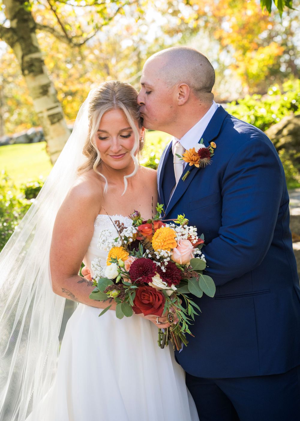 Groom in navy suit kisses bride wearing white gown and veil holding autumn bouquet outdoors.