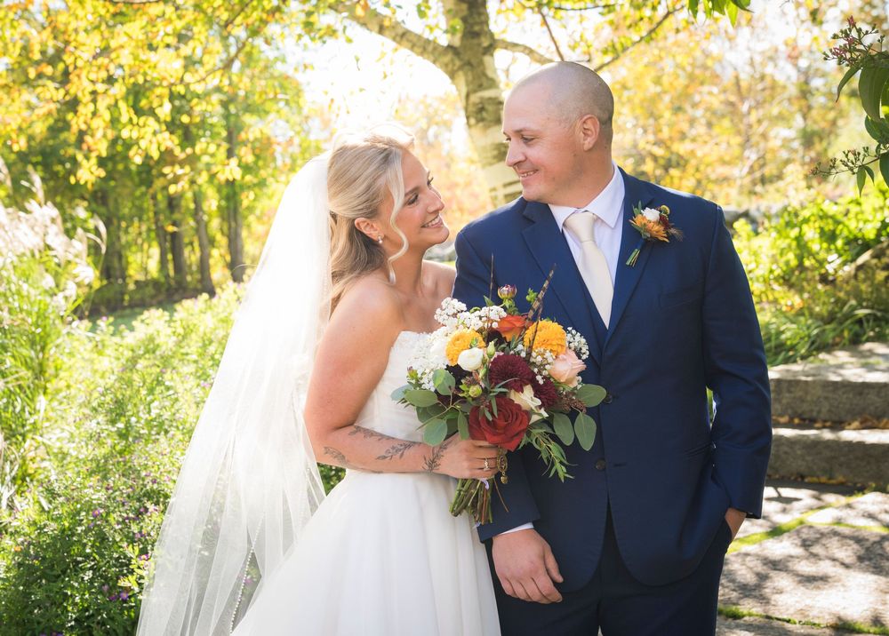 Bride and groom share a romantic glance outdoors, surrounded by golden autumn foliage, bride holding a colorful bouquet.