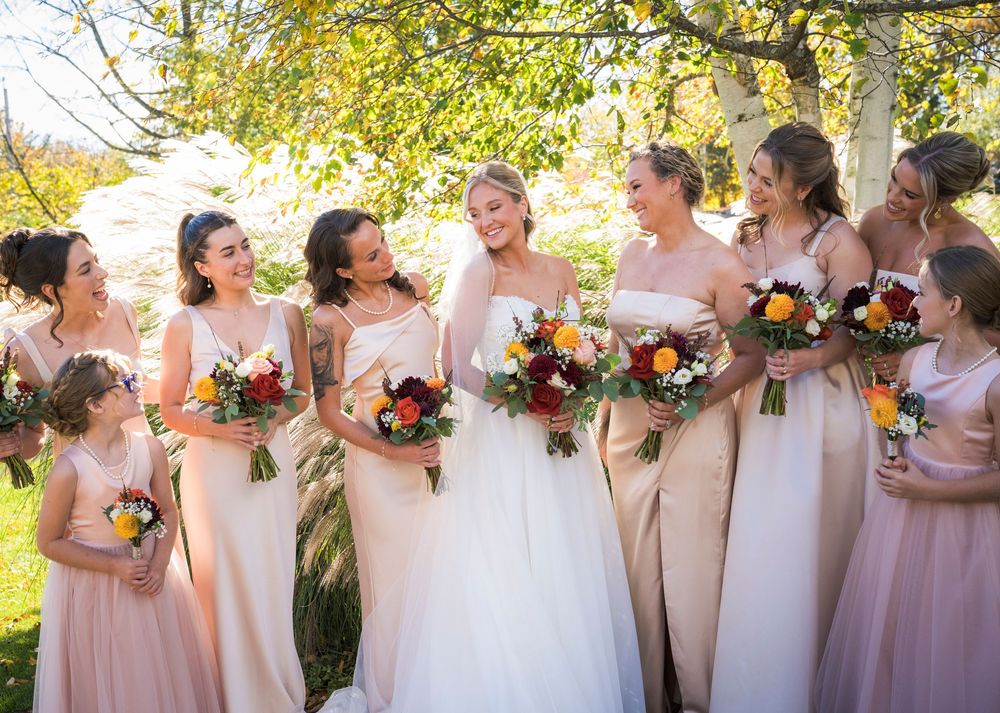 Bride with bridesmaids in blush gowns holding colorful fall bouquets outdoors.