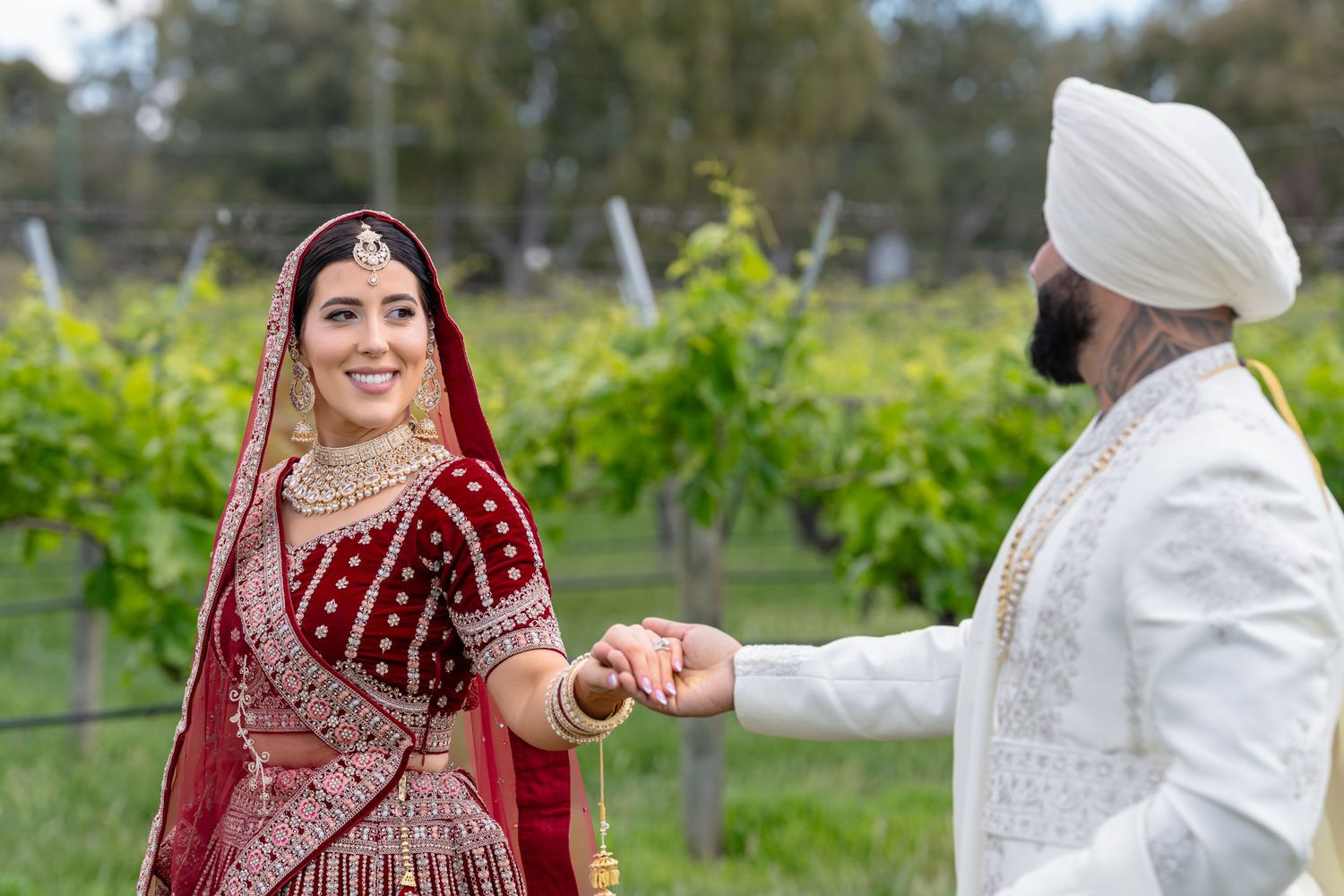 Sikh bride in red lehenga and groom in white sherwani holding hands at a vineyard wedding.