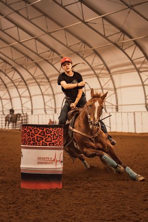 A young barrel racer on a chestnut horse rounds a decorated barrel at a Hearts and Hoofbeats indoor rodeo event.
