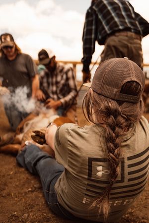 Farmers branding cattle on a ranch, with a woman in an Under Armour shirt holding a calf on the ground.