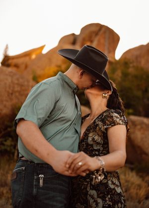 Couple sharing a romantic kiss outdoors near rustic rock formations, man in cowboy hat and green shirt, woman in floral dress.