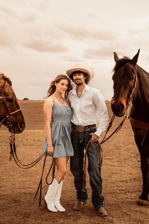 A couple in western wear poses with two brown horses on a dry ranch field under a dramatic cloudy sky.