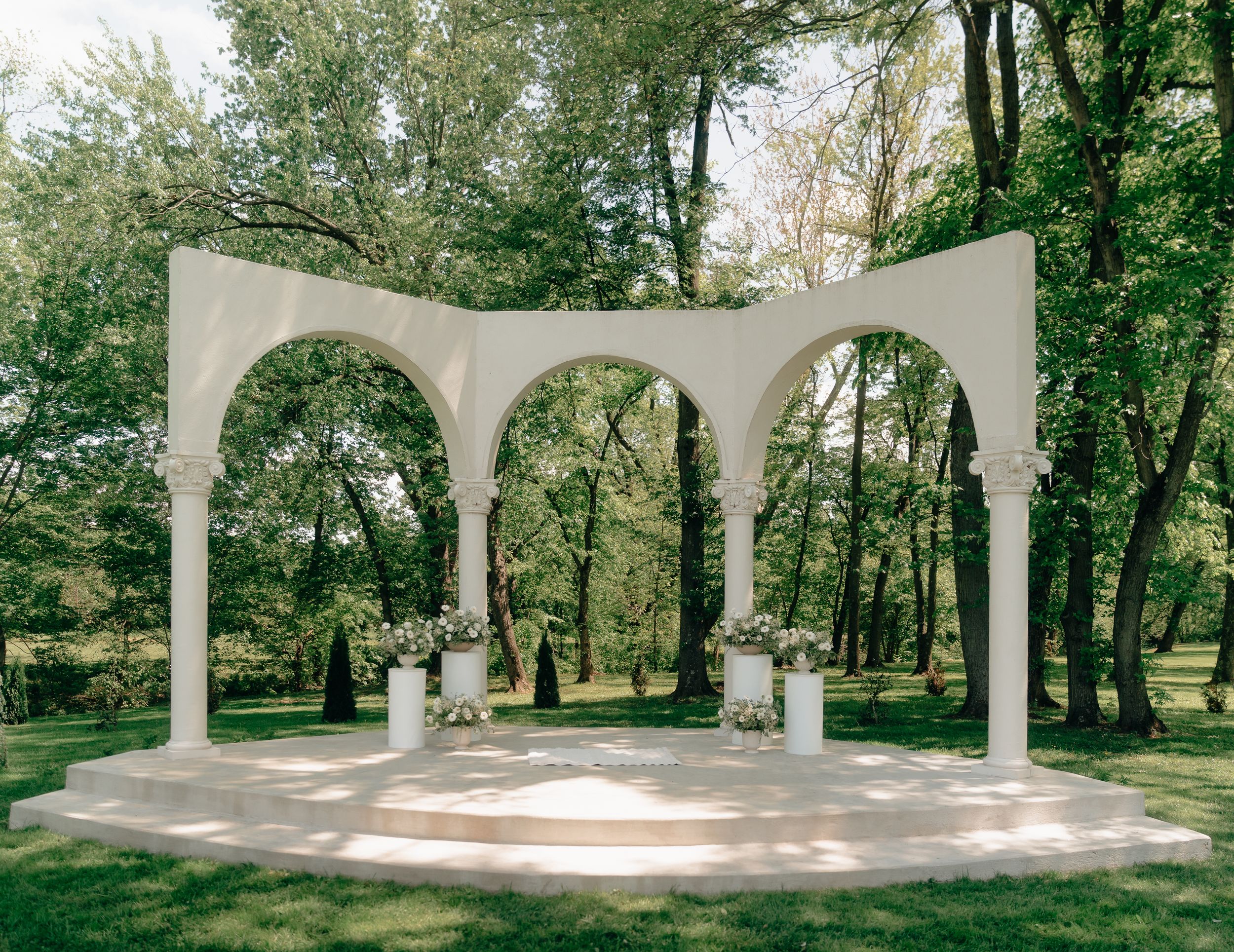 Elegant white arched wedding ceremony structure with columns surrounded by lush green trees in an outdoor garden setting.