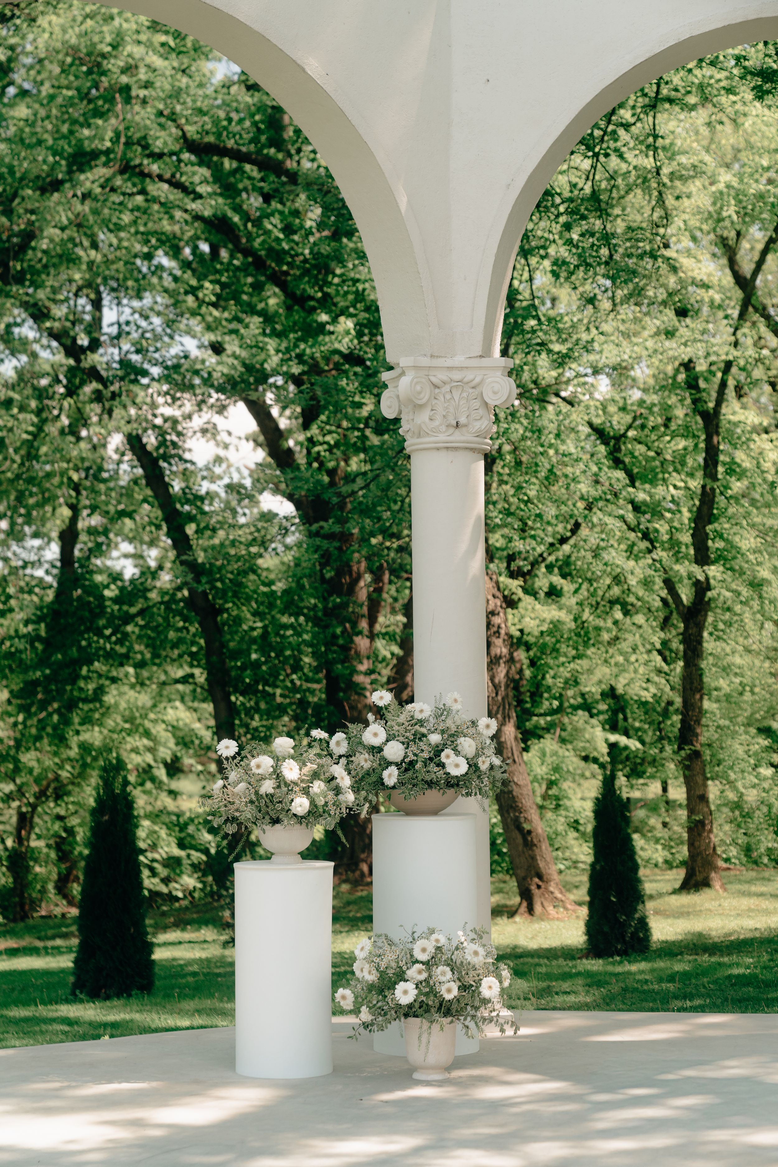 White floral arrangements on pedestals beside elegant arched colonnade in lush green garden wedding venue.