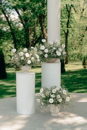 Elegant outdoor wedding decor with white flower arrangements in vases on cylindrical pedestals surrounded by lush greenery.