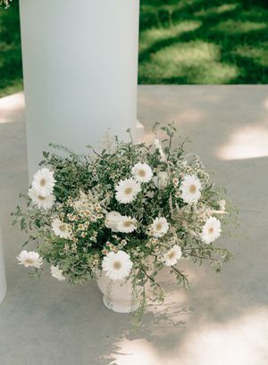 White gerbera daisies and greenery arrangement in a white pot at the base of a column for a wedding ceremony.