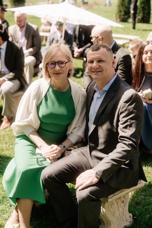 A smiling couple sits together outdoors at a garden event, woman in green dress with glasses, man in dark suit.