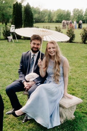 A smiling couple sits on a stone bench outdoors at a wedding, the woman in a blue dress holding a white parasol.