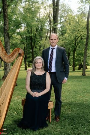 A woman in a black gown sits beside a harp while a man in a suit stands beside her outdoors.
