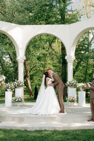 Bride and groom share first kiss under white arched gazebo adorned with floral arrangements in lush garden wedding ceremony.